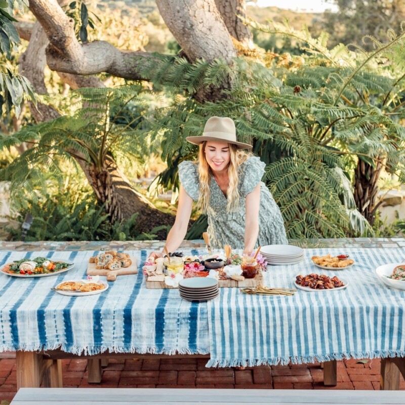 Blonde woman wearing sun hat setting out food dishes on outdoor dining table.