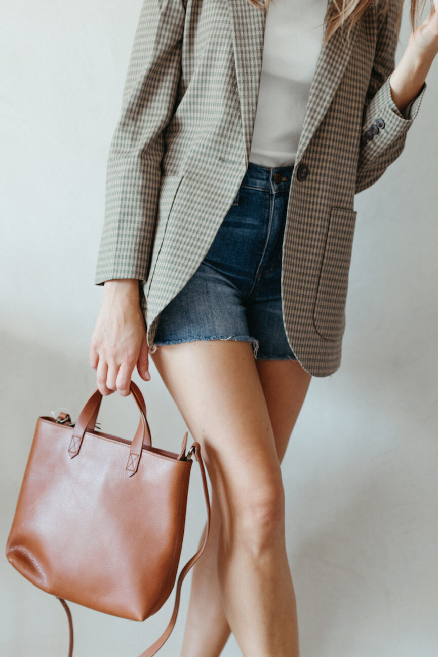 Woman wearing blazer with denim shorts and carrying brown leather handbag.